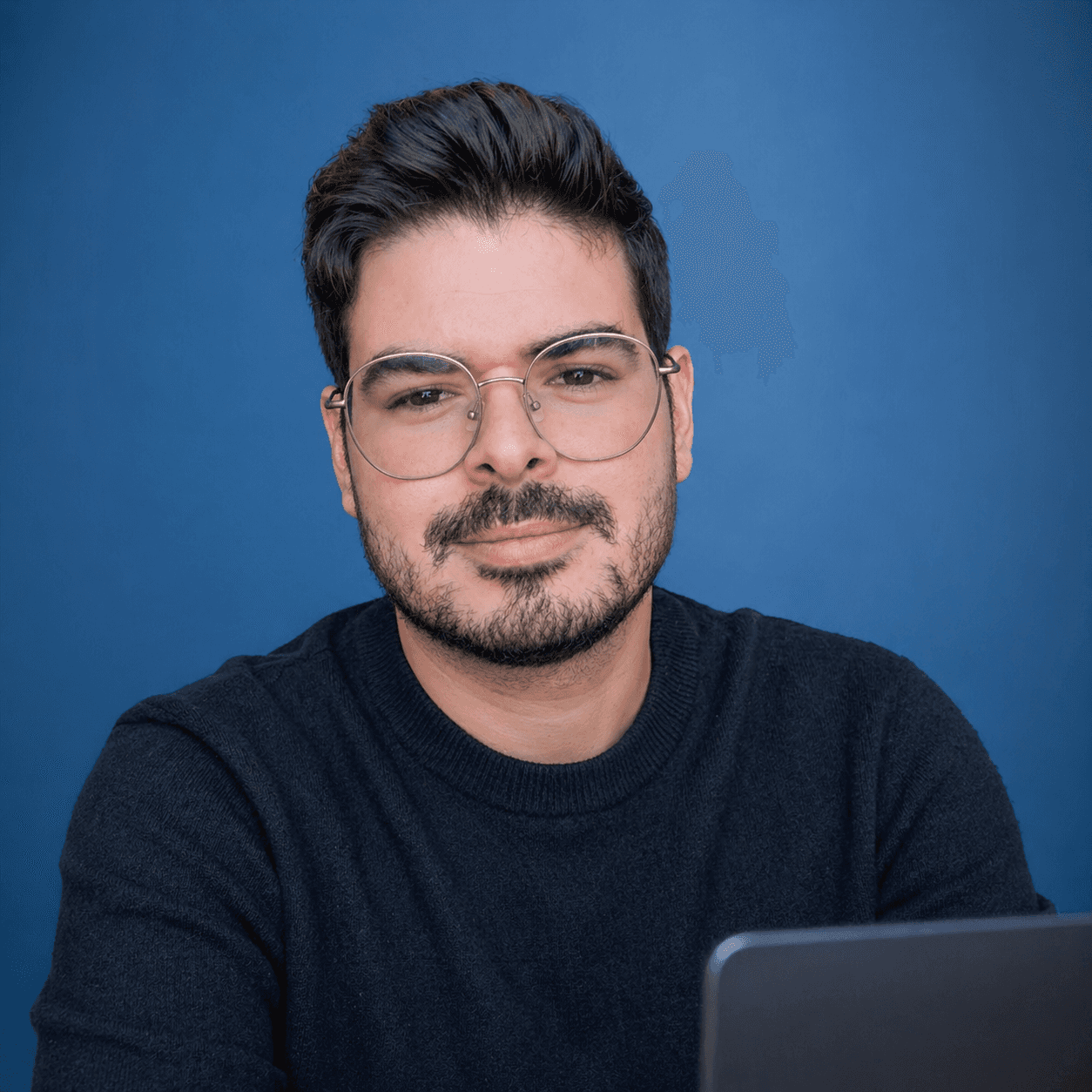 Bearded man with glasses wearing a black sweater, sitting behind a laptop against blue background.